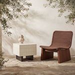 Outdoor setting with side table next to woven brown lounge chair on stone floor under olive tree branches