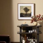 Framed painting displayed on beige wall above dark shelving unit with glassware and decor