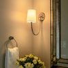 Sconce lit on beige wall next to floral vase and towel ring under large framed mirror in warm light