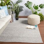 Rug styled in an indoor or patio setting with wicker sofa, beige ottoman, and green plants on wood floor