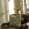 Pillow on brown armchair with floral wallpaper in background and blurred greenery in foreground