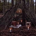 Multiple storm lanterns lit inside a rustic tepee made of tree branches in woodland setting at dusk