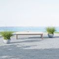 Bench shown outdoors near ocean with taupe cushion and gravel ground, flanked by two potted grasses