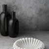 Side view of white marble ruffled bowl with sculptural edge on gray surface with black decorative vases in background