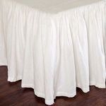 Close-up of gathered cream linen fabric texture and stitching on bed skirt corner over dark hardwood floor