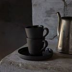 Two black coffee cups stacked on matching saucer with dark rustic background and vintage metal kettle