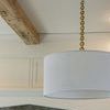 White pleated pendant shade suspended from brass chain against white ceiling and walls with exposed wood beam
