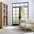 Retreat Pole Rattan End Table placed beside cream upholstered armchair near large window with black frame.