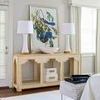 Console shown in bedroom setting with white lamps, artwork, stacked boxes, and ottoman nearby