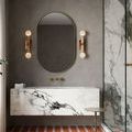 Beatrix Bath Wall Sconce mounted beside oval mirror in modern bathroom with marble and brass fixtures