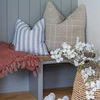 Two plaid pillows styled on a bench with a blue striped pillow, red throw, wicker basket, and white flowers