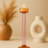 Single glass candle holder lit with flame, styled on beige surface with blurred ceramic decor background