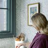 Stillness with Citrus painting hanging on patterned wall above kitchen counter with woman arranging fruit basket nearby