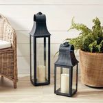 Two Geneva lanterns of different heights styled indoors on a wood floor near wicker furniture and a fern