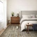 Layla rug under bed in neutral bedroom with wooden dresser, lamp, and slippers on rug edge
