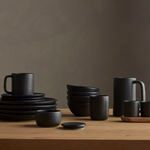 Matte black ceramic tableware set including plates, bowls, mugs, and a pitcher on wooden surface