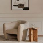 Painting hung centered on a beige wall over a curved beige armchair with a wooden side table