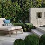 Laguna Double Chaise placed outdoors on concrete patio with greenery and white textured wall background
