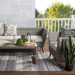 Outdoor porch scene with plants and gray woven furniture placed on trellis-patterned, fringed gray rug