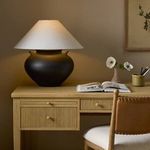 Civita table lamp illuminated on wood desk next to open book, ceramic vase with flowers, and leather chair in warm-toned room