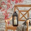 Close-up side view of Antoinne Outdoor Lantern on wooden chair with grey throw and stone wall background