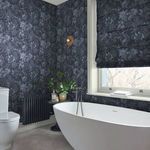 Wallpaper applied in a bathroom corner surrounding a freestanding white bathtub and window with matching roman shade