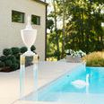 Clear acrylic pedestal used outdoors next to pool, with white urn on top and greenery background