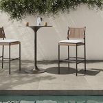 Outdoor scene with two stools and metal pedestal table by tiled pool area and white wall with greenery