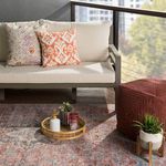 Indoor/outdoor rug placed under red corduroy pouf next to green potted plant on wood outdoor flooring