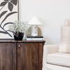 Bond lamp on wooden credenza beside floral painting, large vase with flowers, and beige couch pillow