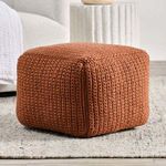 Rust pouf placed on a neutral rug beside a cream chair and textured white pedestal in a room setting