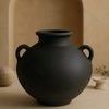Black terracotta vase placed on beige surface with arch backdrop and decorative objects in soft warm lighting
