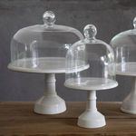 Set of three white footed plates with varying sizes of clear glass domes on wooden table against gray wall