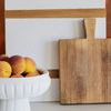 Close-up of white pedestal bowl filled with peaches next to wooden and white cutting boards on marble countertop