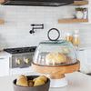 Bread loaf inside glass dome of cake stand on a marble island next to dark brown bowl of pears with kitchen stove in backg...