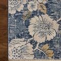 Corner of rug on wood floor showing cream floral pattern, denim background, and beige fringe edge