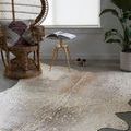 Bryce rug displayed flat on a gray floor beneath a wicker chair and a small gold side table with an open book