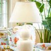 Fulton Vase Lamp lit on a glass side table in a sunlit room with floral upholstered chair and large green plant