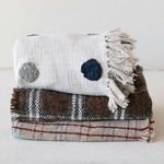 Side view of a stack of folded throws featuring the plaid Fireside Fringe with other textured blankets