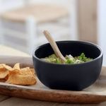 Side view of seed grey bowl filled with guacamole and wooden spoon on wooden tray with chips