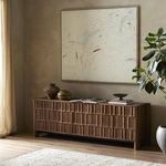 Painting displayed in a warm living room setting above carved wood sideboard with plants and decor