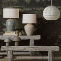 Styled scene with Mimi Ivory Table Lamp on rustic bench against a dark nature mural wall with other lamps and natural decor
