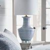Lamp shown in bedroom setting on a nightstand with bed pillows and natural light from nearby windows