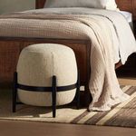 Sia Ottoman placed on rug in a bedroom with beige textured bedding and wooden bed frame