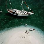 Closer aerial crop focusing on the yacht deck, colorful signal flags, people swimming, and the small sandy island below
