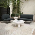Outdoor scene of Salma chair and loveseat on beige rug next to round white coffee table and greenery backdrop