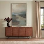 Painting hung above rattan wood credenza in neutral-toned living room with light drapes and window