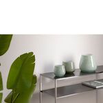 Three different sizes of greenish glass lantern vases styled on metal shelf with green plant nearby