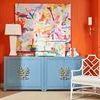 White rattan bench in vibrant room with orange walls, blue cabinet, and colorful abstract paintings