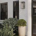 Outdoor wall sconce installed on exterior white brick wall above greenery and planter near black-framed windows and door